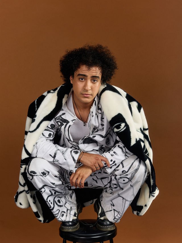 Adam El Mouna accroupi sur un tabouret, vêtu d'un ensemble peint à la main noir et blanc